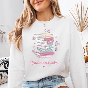 Puede incluir: Sudadera blanca con una pila de libros impresa en la parte delantera. Los libros tienen cubiertas rosas y azules con títulos como "Red, White & Royal Blue" y "C.L.U.P.A.H.Y.A." El texto "Read more books" está impreso debajo de la pila de libros.