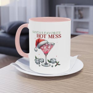 Könnte beinhalten: Eine weiße Keramiktasse mit rosafarbenem Rand und Henkel. Die Tasse zeigt einen roten Weihnachtsmann-Hut mit einem rosafarbenen Cocktailglas, gefüllt mit Eis und einem rosafarbenen Getränk. Der Text auf der Tasse lautet "Santa's Favorite Hot Mess".