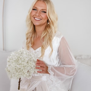 May include: A bride wearing a white satin and lace robe holds a bouquet of white hydrangeas.