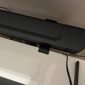 Op de afbeelding: Een zwarte soundbar met volumeknoppen en een aan/uit-knop. De soundbar is gemonteerd op een wit oppervlak.