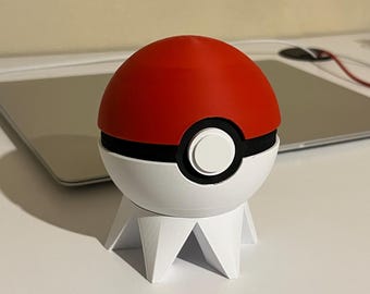 May include: A 3D printed Pokeball with a red top, black band, and white bottom. The Pokeball is sitting on a white, 3D printed stand with three legs.
