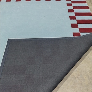 May include: A rectangular area rug featuring a light blue center and a border composed of alternating dark red and light blue rectangular blocks. One corner of the rug is folded over to reveal a black, textured, woven backing material.