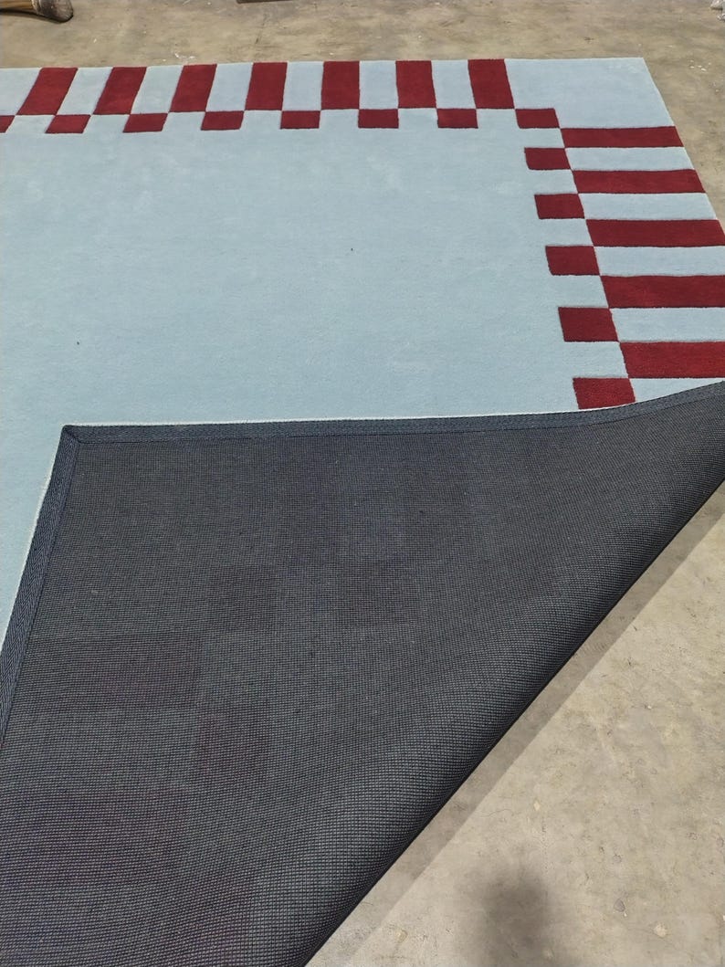 May include: A rectangular area rug featuring a light blue center and a border composed of alternating dark red and light blue rectangular blocks. One corner of the rug is folded over to reveal a black, textured, woven backing material.