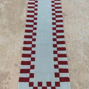 May include: A rectangular runner rug featuring a solid light blue center panel surrounded by a decorative border of alternating dark red and light blue square blocks, creating a geometric pattern along the entire perimeter of the floor covering.
