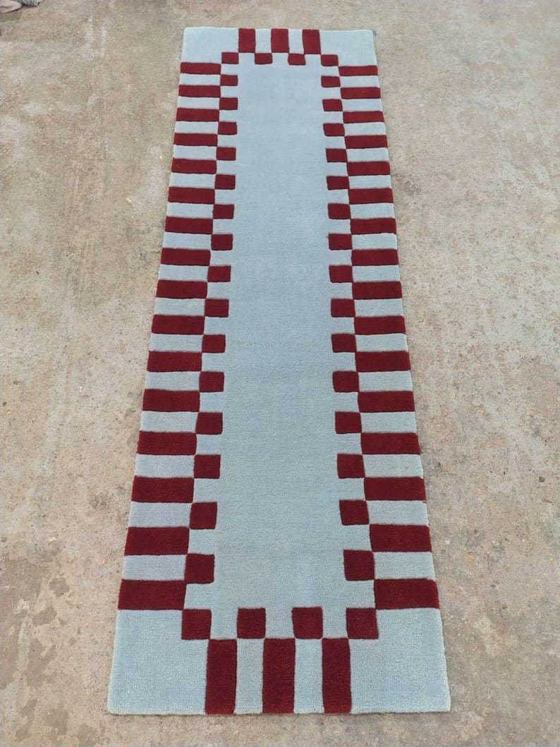 May include: A rectangular runner rug featuring a solid light blue center panel surrounded by a decorative border of alternating dark red and light blue square blocks, creating a geometric pattern along the entire perimeter of the floor covering.