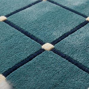 May include: Close-up of a teal rug with a diamond pattern. The diamonds are outlined in navy blue and have small, off-white squares at the intersections. The rug has a soft, textured appearance.