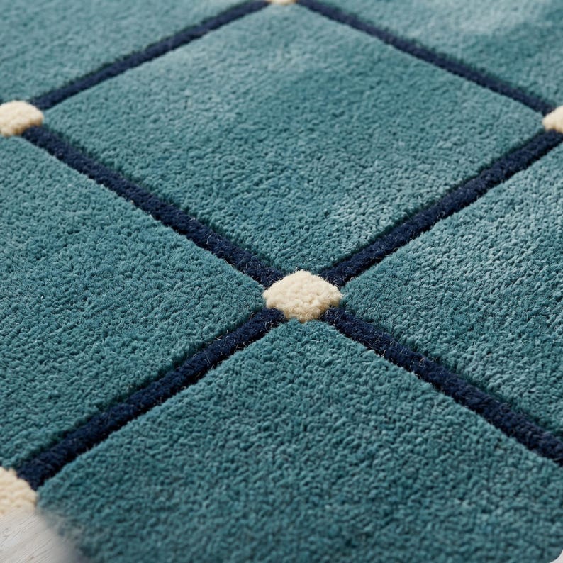 May include: Close-up of a teal rug with a diamond pattern. The diamonds are outlined in navy blue and have small, off-white squares at the intersections. The rug has a soft, textured appearance.