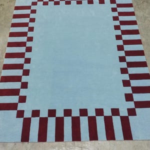 May include: A rectangular area rug featuring a solid light blue center field surrounded by a decorative border composed of alternating dark red and light blue rectangular blocks, creating a structured geometric pattern across the entire surface.