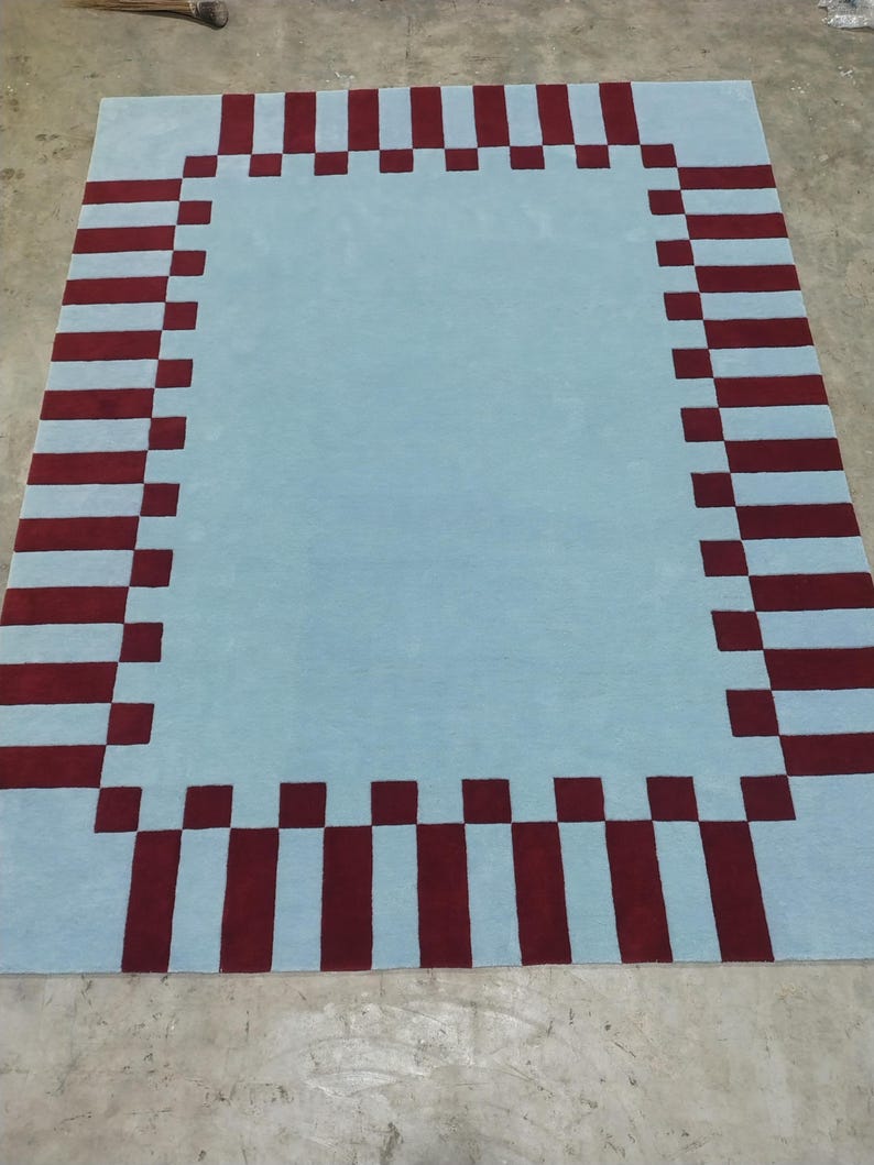 May include: A rectangular area rug featuring a solid light blue center field surrounded by a decorative border composed of alternating dark red and light blue rectangular blocks, creating a structured geometric pattern across the entire surface.