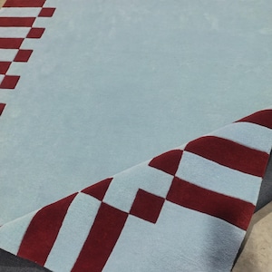 May include: A large area rug featuring a light blue base color with a dark red geometric border pattern. The rug has a tufted texture and one corner is folded over to reveal the dark gray backing material underneath the primary surface.
