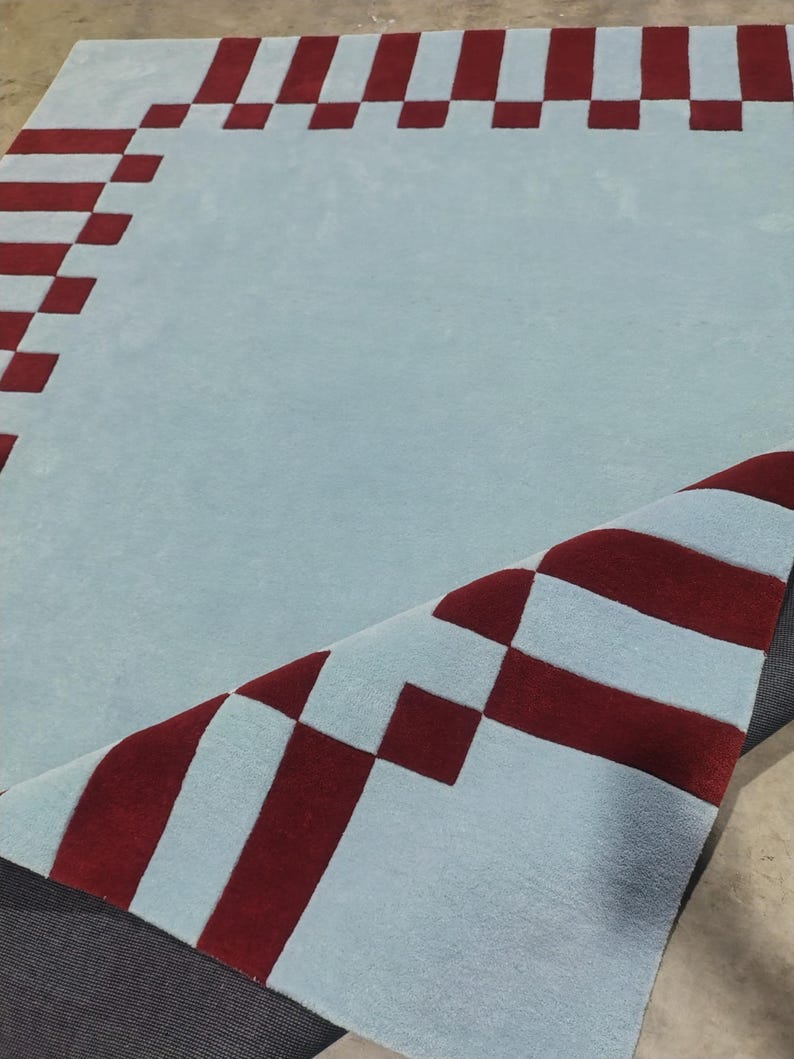 May include: A large area rug featuring a light blue base color with a dark red geometric border pattern. The rug has a tufted texture and one corner is folded over to reveal the dark gray backing material underneath the primary surface.