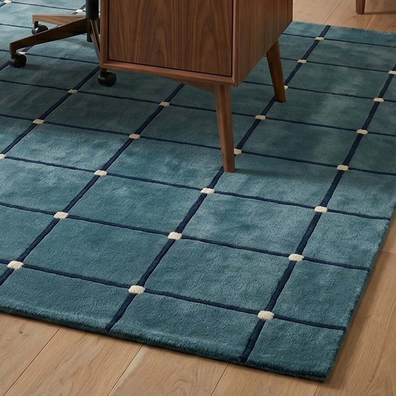 May include: Teal area rug with a grid pattern. The rug features a teal background with a dark blue grid design and small off-white squares at the intersections. A wooden desk and chair are partially visible in the background.
