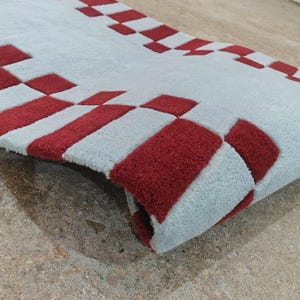 May include: A rectangular area rug featuring a light blue base color with a repeating pattern of deep red squares arranged along the edges. The rug has a thick, tufted texture and is shown partially folded to reveal the underside and pile depth.
