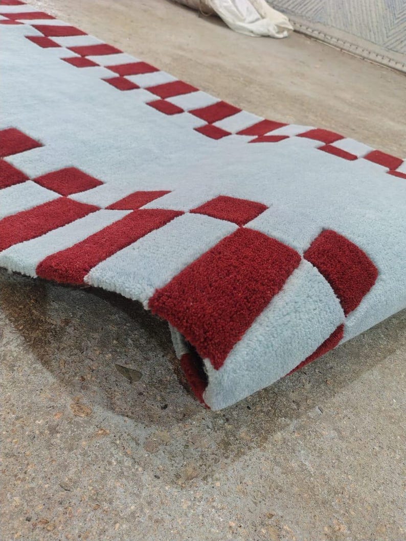 May include: A rectangular area rug featuring a light blue base color with a repeating pattern of deep red squares arranged along the edges. The rug has a thick, tufted texture and is shown partially folded to reveal the underside and pile depth.