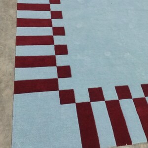 May include: A large area rug featuring a solid light blue center field surrounded by a wide border. The border consists of alternating dark red rectangular blocks and light blue squares, creating a geometric pattern along the edges of the rug.