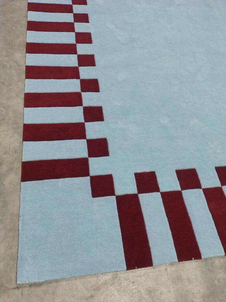 May include: A large area rug featuring a solid light blue center field surrounded by a wide border. The border consists of alternating dark red rectangular blocks and light blue squares, creating a geometric pattern along the edges of the rug.