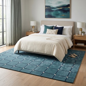 May include: A bedroom scene featuring a teal blue rug with a grid pattern, a bed with a cream-colored comforter, and decorative pillows. Two bedside tables with lamps flank the bed. A painting hangs above the bed, and the room has hardwood floors and curtains.