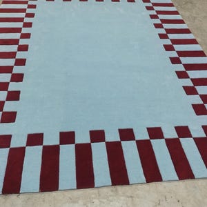May include: A rectangular area rug featuring a solid light blue center field surrounded by a wide border. The border consists of alternating dark red rectangular blocks and light blue squares, creating a repeating geometric pattern around the edge.