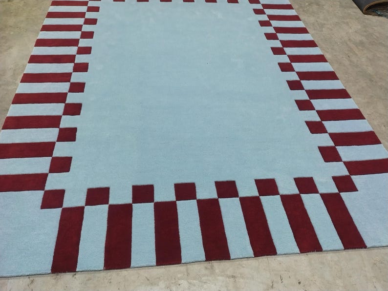 May include: A rectangular area rug featuring a solid light blue center field surrounded by a wide border. The border consists of alternating dark red rectangular blocks and light blue squares, creating a repeating geometric pattern around the edge.
