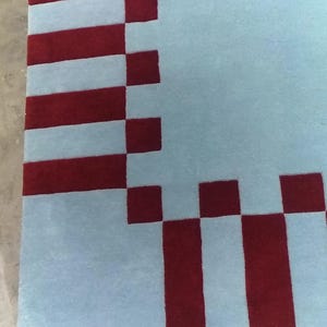 May include: A rectangular area rug featuring a light blue field bordered by a geometric pattern of alternating dark red and light blue rectangular blocks. The design creates a structured frame around the central light blue section of the rug.