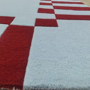 May include: A rectangular area rug featuring a light blue base color with a geometric border pattern composed of deep red rectangular blocks. The rug has a thick, tufted pile texture that creates a distinct contrast between the red and blue sections.