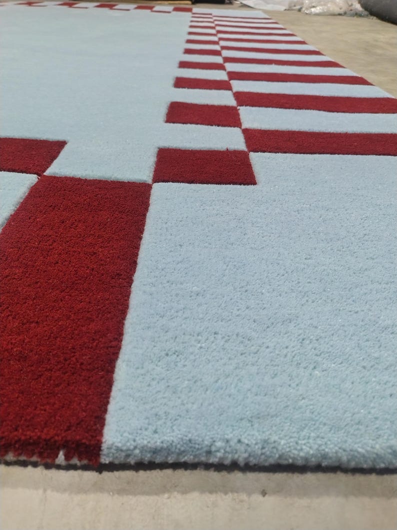 May include: A rectangular area rug featuring a light blue base color with a geometric border pattern composed of deep red rectangular blocks. The rug has a thick, tufted pile texture that creates a distinct contrast between the red and blue sections.