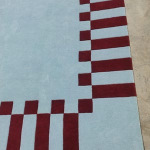 May include: A rectangular area rug featuring a solid light blue center field bordered by a repeating pattern of deep red squares and vertical rectangular stripes that frame the perimeter of the light blue surface in a geometric arrangement.
