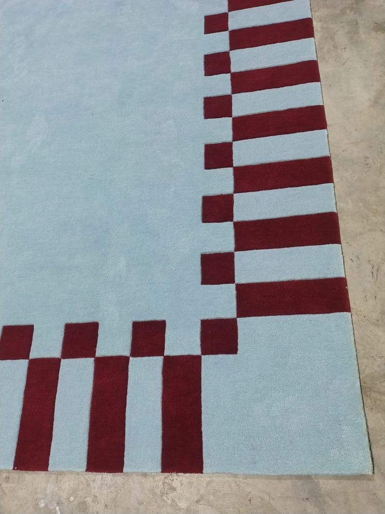 May include: A rectangular area rug featuring a solid light blue center field bordered by a repeating pattern of deep red squares and vertical rectangular stripes that frame the perimeter of the light blue surface in a geometric arrangement.