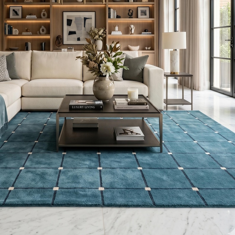 May include: A teal blue area rug with a grid pattern of white squares and dark blue lines. The rug is in a living room setting with a white sofa, dark wood coffee table, and a bookshelf. The coffee table has a book that says "LUXURY LIVING".