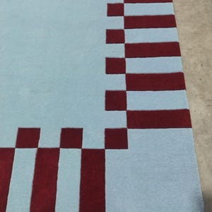 May include: A rectangular area rug featuring a solid light blue center field surrounded by a border of alternating dark red and light blue rectangular blocks. The rug has a flat, uniform texture and a clean, geometric pattern along the edges.