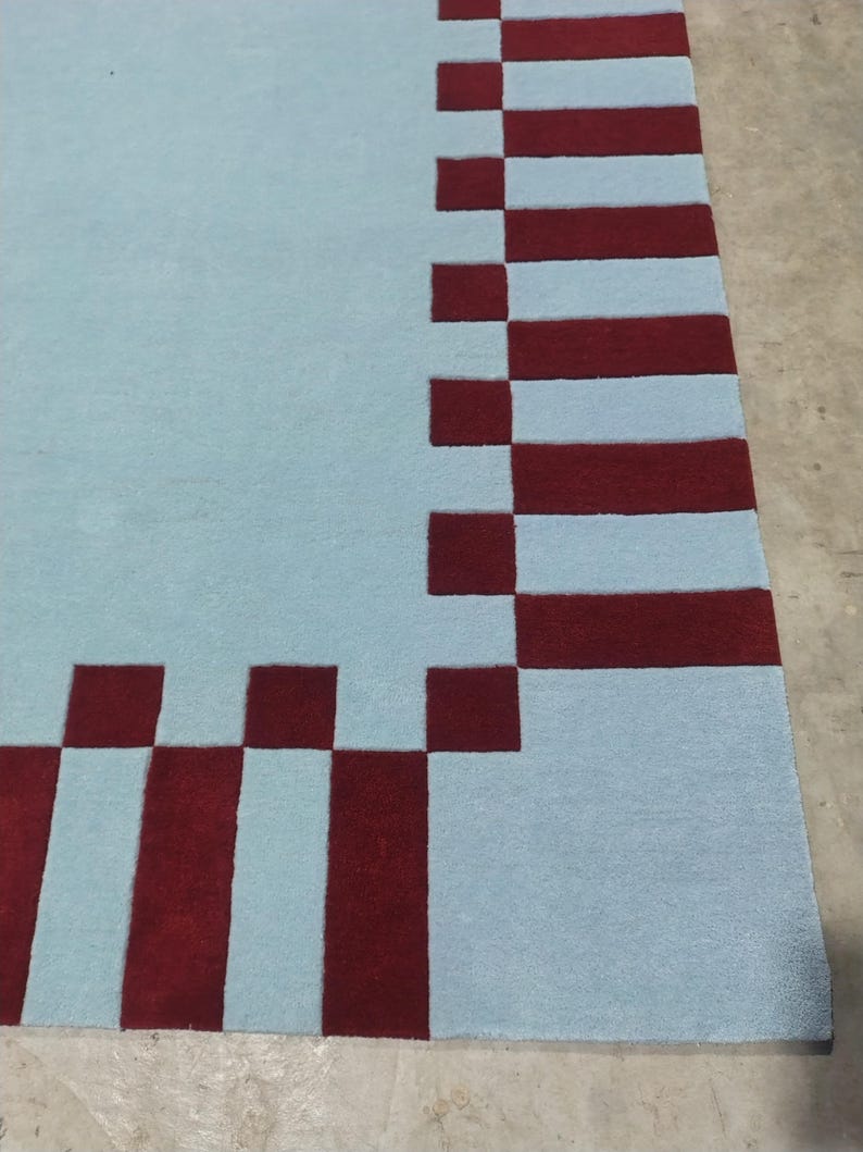 May include: A rectangular area rug featuring a solid light blue center field surrounded by a border of alternating dark red and light blue rectangular blocks. The rug has a flat, uniform texture and a clean, geometric pattern along the edges.