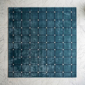May include: A square teal rug with a grid pattern. Dark navy lines create a grid, with small, square, off-white accents at each intersection. The rug has a soft, textured appearance, suitable for a living room or bedroom.