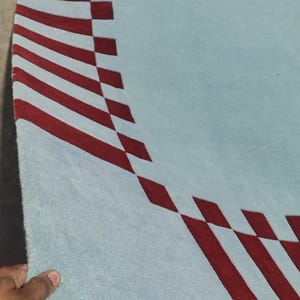 May include: A light blue area rug featuring a curved border pattern composed of deep red rectangular shapes arranged in a repeating sequence along the edge, creating a distinct contrast against the solid light blue field of the textile surface.