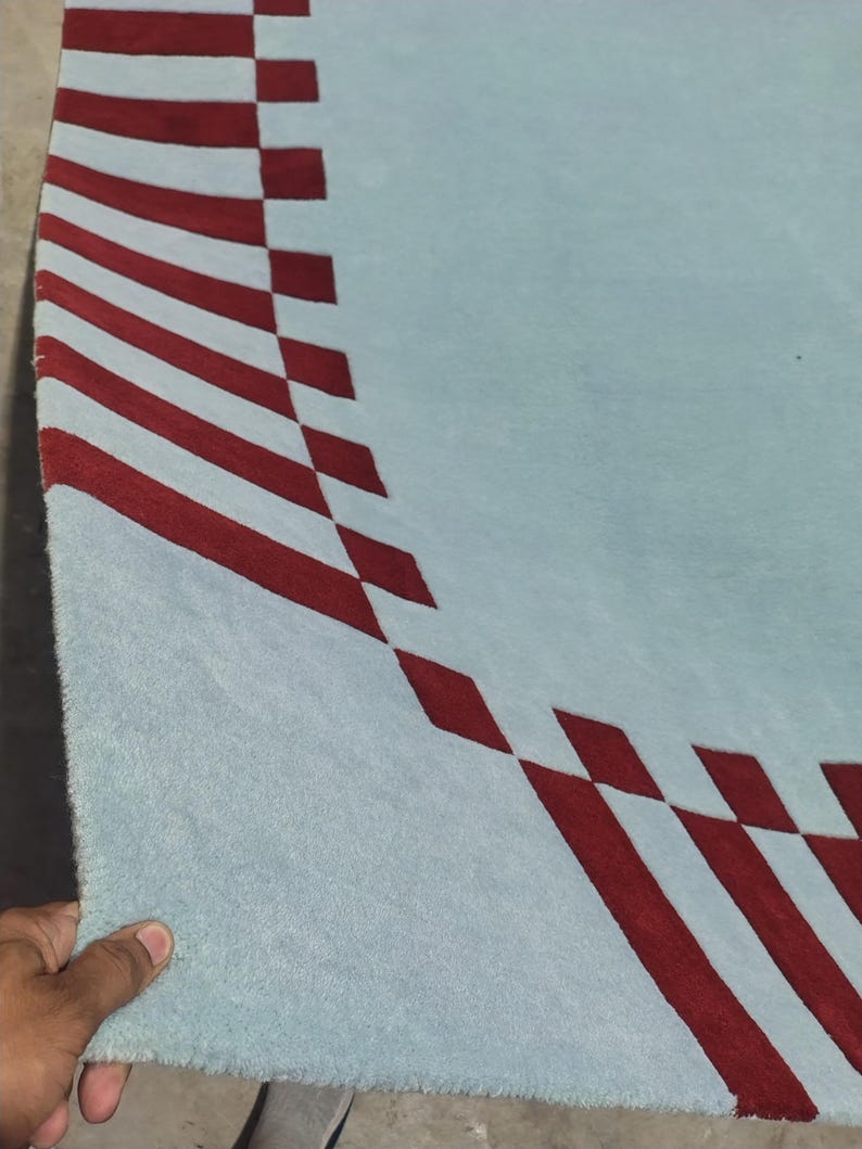 May include: A light blue area rug featuring a curved border pattern composed of deep red rectangular shapes arranged in a repeating sequence along the edge, creating a distinct contrast against the solid light blue field of the textile surface.