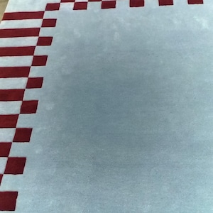 May include: A rectangular area rug featuring a solid light blue center field surrounded by a border of alternating dark red and light blue square blocks. The rug has a dense, tufted texture and a clean, geometric pattern along its outer edges.