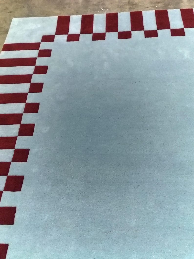 May include: A rectangular area rug featuring a solid light blue center field surrounded by a border of alternating dark red and light blue square blocks. The rug has a dense, tufted texture and a clean, geometric pattern along its outer edges.