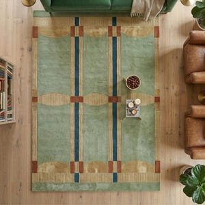 Puede incluir: Vista a&eacute;rea de una alfombra rectangular con un campo verde claro. La alfombra presenta rayas verticales azul marino y formas geom&eacute;tricas decorativas de color beige y &oacute;xido. La alfombra est&aacute; colocada sobre un suelo de madera, con un sof&aacute; verde y sillones de cuero visibles.