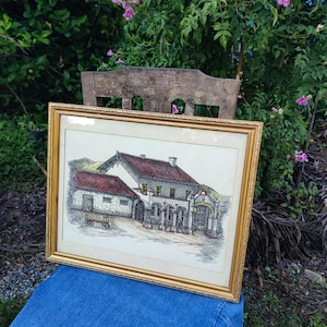 May include: A framed black and white drawing of a building with a red tile roof. The building has a large arched entrance and several windows. The drawing is set against a background of hills and trees.