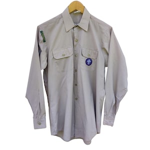 May include: A beige long-sleeved button-up shirt with two chest pockets and a scout emblem on the left pocket.
