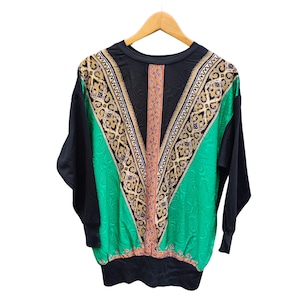 Puede incluir: Top negro de manga larga con un diseño único. El top presenta un panel en forma de V en verde y dorado con un patrón intrincado. Las mangas y la espalda son de color negro liso. El top está colgado en una percha de madera.