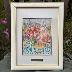 Op de afbeelding: Een ingelijst bloemstilleven schilderij. Het kunstwerk toont een kleurrijk boeket in een gele vaas, tegen een blauw-wit gestreepte achtergrond. De lijst is wit met een gouden binnenrand. De naam van de kunstenaar, Leslie Sayour, is weergegeven.