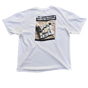 May include: A white short-sleeved t-shirt featuring a graphic on the back. The graphic shows a grid of square images and the text "Nobody puts Windows NT together like Intergraph." The shirt has a crew neckline and a standard fit design.