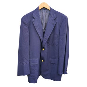 Puede incluir: Un blazer azul marino con botones dorados, colgado de una percha de madera. El blazer tiene dos bolsillos delanteros y un bolsillo en el pecho. La tela parece ser tejida.