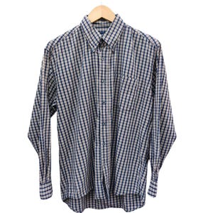 Puede incluir: Camisa abotonada de manga larga con un clásico estampado de cuadros en azul marino, blanco y beige. La camisa presenta un cuello abotonado, un bolsillo en el pecho y está colgada de una percha de madera sobre un fondo blanco.