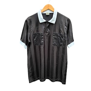 May include: Black and gray striped polo shirt with a light blue collar and cuffs. The shirt has two chest pockets and a three-button placket. The shirt is hanging on a wooden hanger.