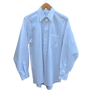 Puede incluir: Una camisa blanca de manga larga con bolsillo en el pecho y cuello clásico, colgada de una percha de madera. El nombre de la marca "fabrique" es visible en el cuello. La camisa está abotonada.