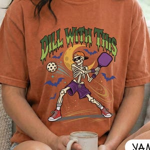 May include: A rust-colored short-sleeve t-shirt featuring a graphic of a skeleton wearing a cap and sunglasses, holding a purple pickleball paddle. The text "DILL WITH THIS" appears in green above the skeleton, surrounded by bats and a yellow moon.