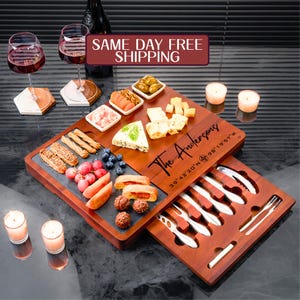 May include: A wooden cheese board with a sliding drawer that holds a set of cheese knives. The board has a slate insert and is engraved with the text "The Andersons" and coordinates. The board is filled with cheese, crackers, grapes, olives, and other snacks.