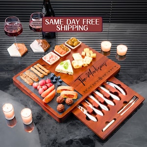 May include: A wooden charcuterie board with a slate insert, filled with various cheeses, fruits, and snacks. Includes a set of serving utensils. The board is personalized with the text "The Madisons" and coordinates. Two wine glasses and candles are also present.
