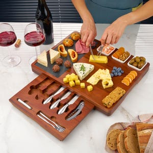 K&ouml;nnte beinhalten: Ein rechteckiges K&auml;sebrett aus Holz mit einer ausziehbaren Schublade, die Servierbesteck aus Edelstahl wie Messer und eine Gabel enth&auml;lt. Das Brett ist mit einer Auswahl an K&auml;se, Crackern, Obst und Fleisch sowie zwei Weingl&auml;sern best&uuml;ckt.